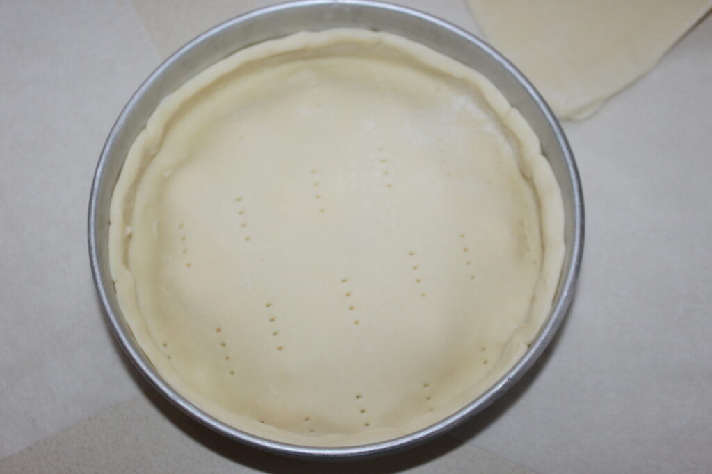 Une croûte à tarte percée de trous dans un moule rond, parfaitement préparée et prête à cuire, repose sur un comptoir blanc. La promesse d'incorporer du chèvre frais délicat laisse entrevoir les saveurs à venir.