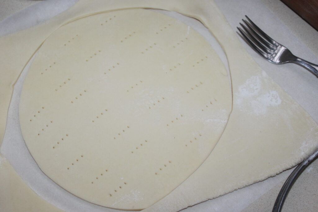 Une pâte feuilletée circulaire non cuite percée de trous de fourchette repose sur du papier sulfurisé, laissant entrevoir une délicieuse tarte tatin en préparation, à côté d'une fourchette sur le comptoir.