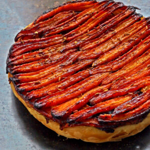 Cette tarte Tatin présente des carottes caramélisées et une touche de chèvre frais, élégamment superposées sur une croûte feuilletée, présentée sur une surface grise.