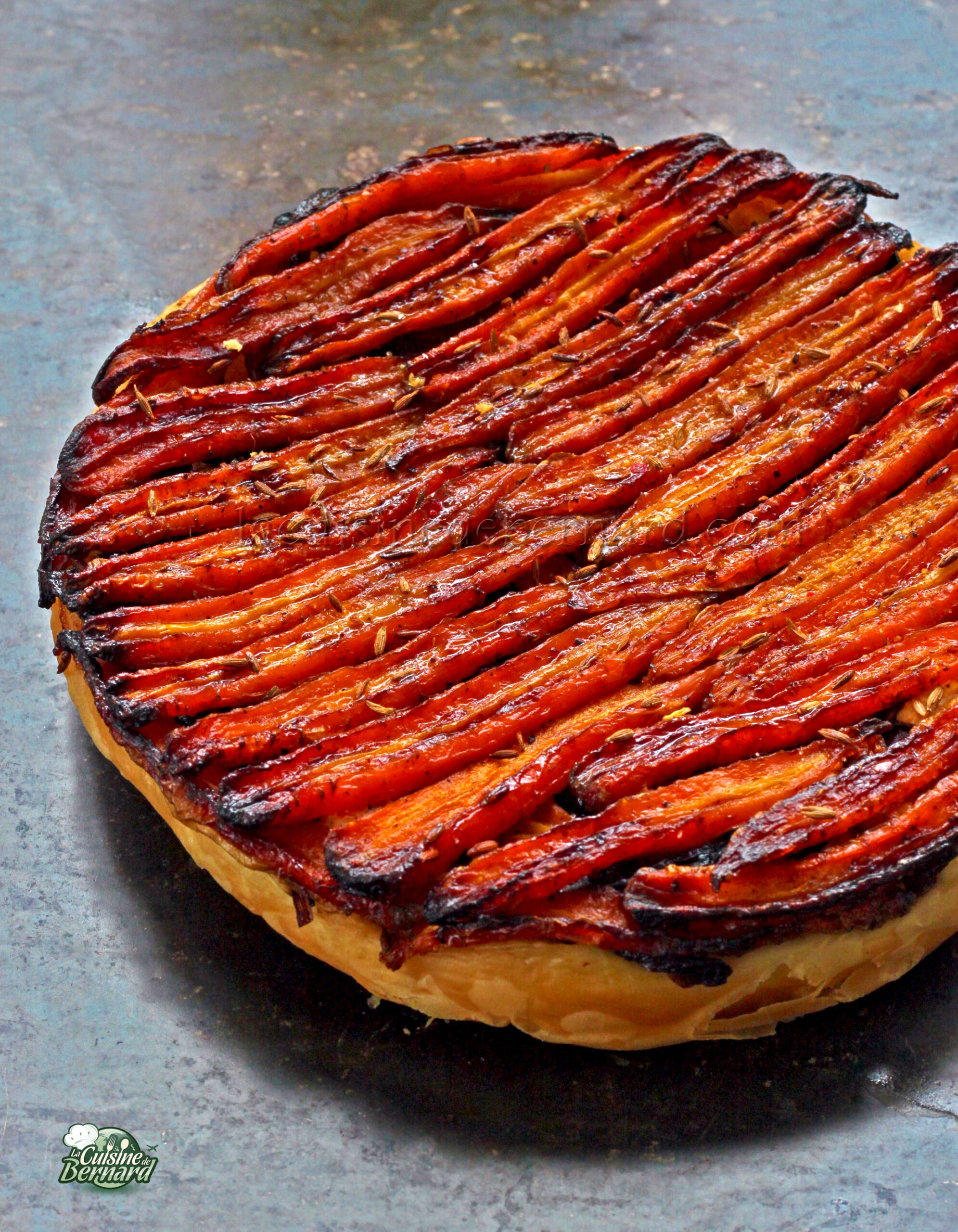 Cette tarte Tatin présente des carottes caramélisées et une touche de chèvre frais, élégamment superposées sur une croûte feuilletée, présentée sur une surface grise.