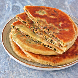 Assiette de pains plats tranchés et farcis, dotés d'une croûte brun doré et d'une garniture végétale visible, notamment de savoureux shitakés, placés sur une surface bleue texturée.