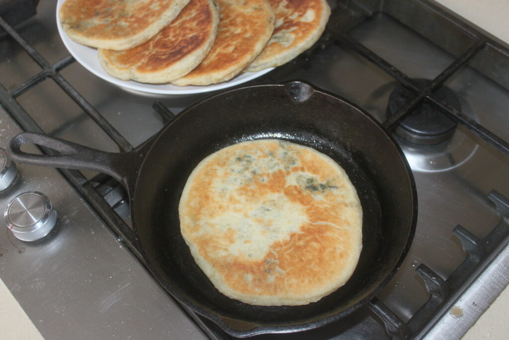 Du pain plat et des galettes grésillent dans une poêle noire sur la cuisinière, tandis que d'autres sont astucieusement empilés sur une assiette blanche en arrière plan, prêts à être associés à de savoureux shitakés.
