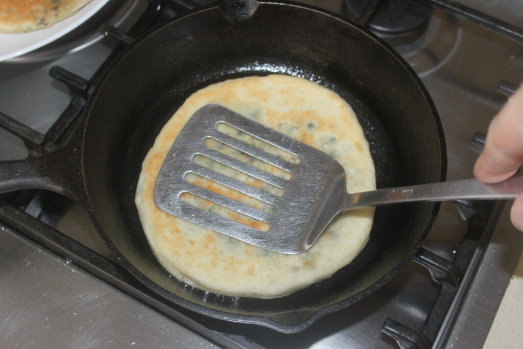 Une personne appuie une spatule sur une galette en train de cuire dans une poêle noire sur une cuisinière.