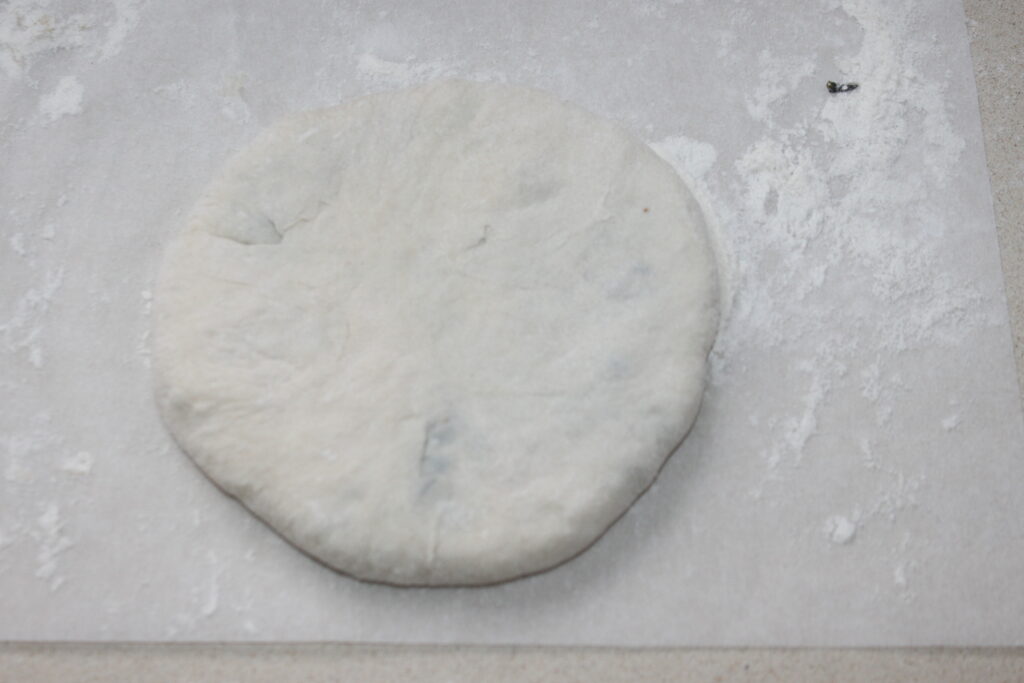 Une boule ronde et aplatie de pâte crue repose sur une feuille de papier sulfurisé, saupoudrée de farine et prête à être transformée en une délicieuse galette.