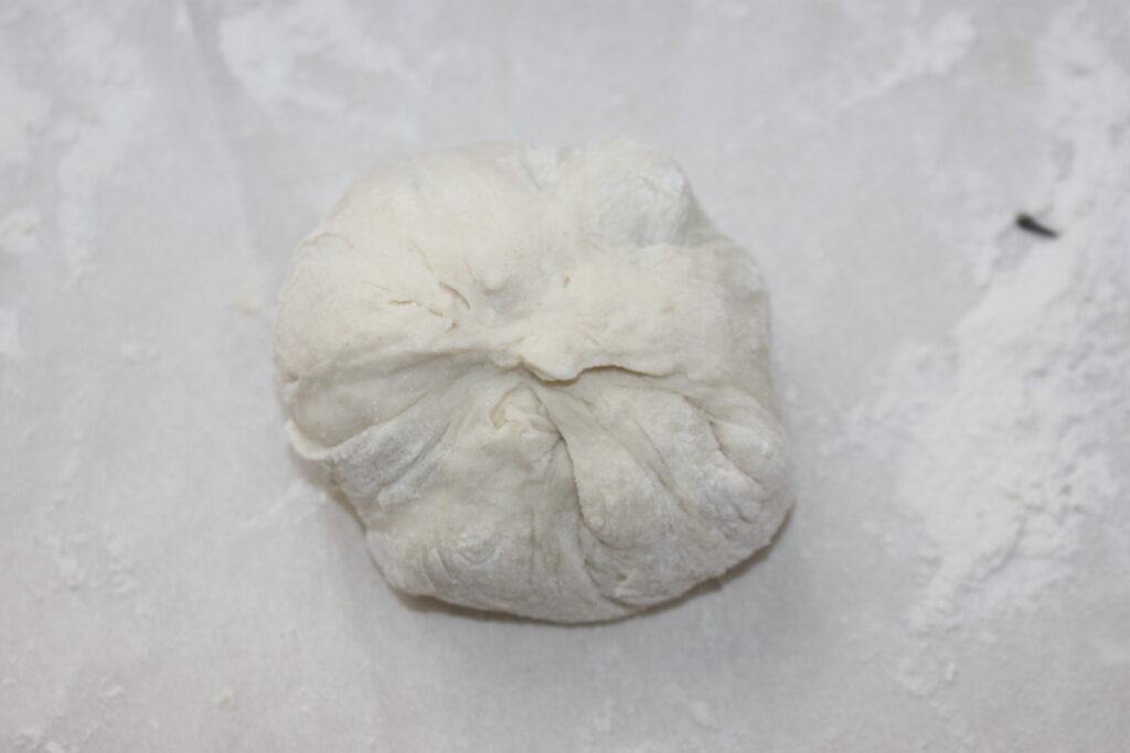 Une boule de pâte crue sur une surface farinée, avec des plis et des replis visibles sur son dessus, prête à être transformée en délicieuses galettes.