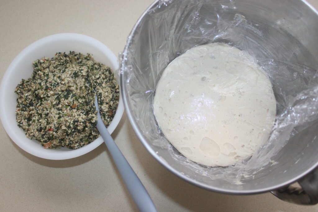 Un bol de shitakés et de légumes est posé à côté d'un saladier contenant de la pâte levée recouverte d'une pellicule plastique. Une cuillère repose dans le mélange de légumes, prêt à être combiné pour des galettes appétissantes.