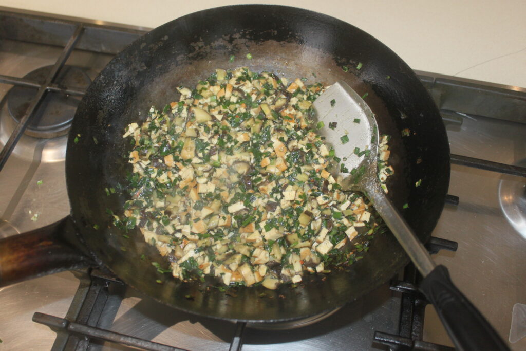 Des légumes hachés et des shitakés sont sautés avec du tofu dans une poêle noire sur la cuisinière, avec une spatule en métal posée à l'intérieur de la poêle.
