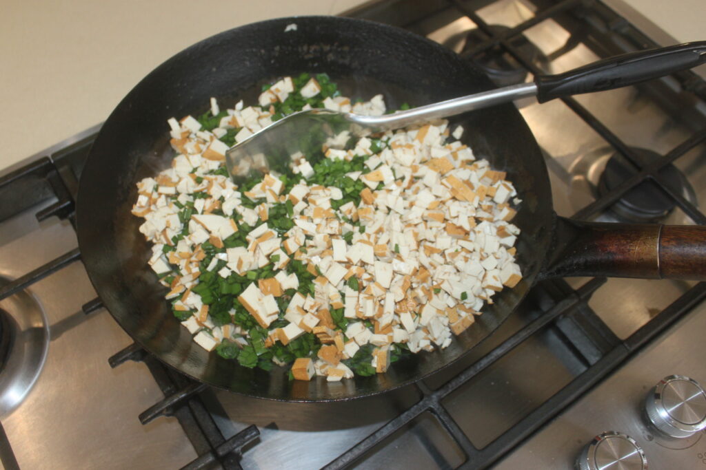 Du tofu coupé en dés et des légumes verts hachés, accompagnés de shitakés tranchés, sont sautés dans une poêle noire sur une cuisinière à gaz avec une spatule en métal.