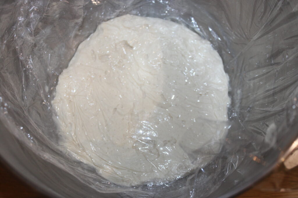 Un bol avec de la pâte à pizza, bientôt garnie de shitakés savoureux, recouvert d'une pellicule plastique transparente.