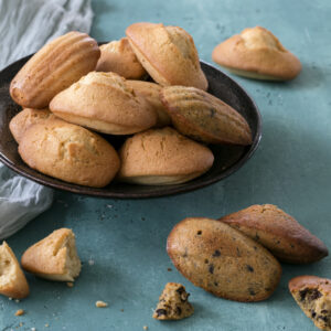 Une assiette de délicieuses pâtisseries françaises repose sur une surface turquoise, avec quelques madeleines éparpillées et partiellement mangées à proximité. Un tissu gris est drapé sur le côté, ajoutant une touche d'élégance à cette scène alléchante.