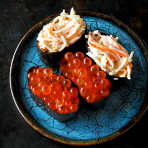 Une assiette bleue avec deux morceaux de sushi garnis de chair de crabe râpée et deux morceaux garnis d'œufs de poisson orange.