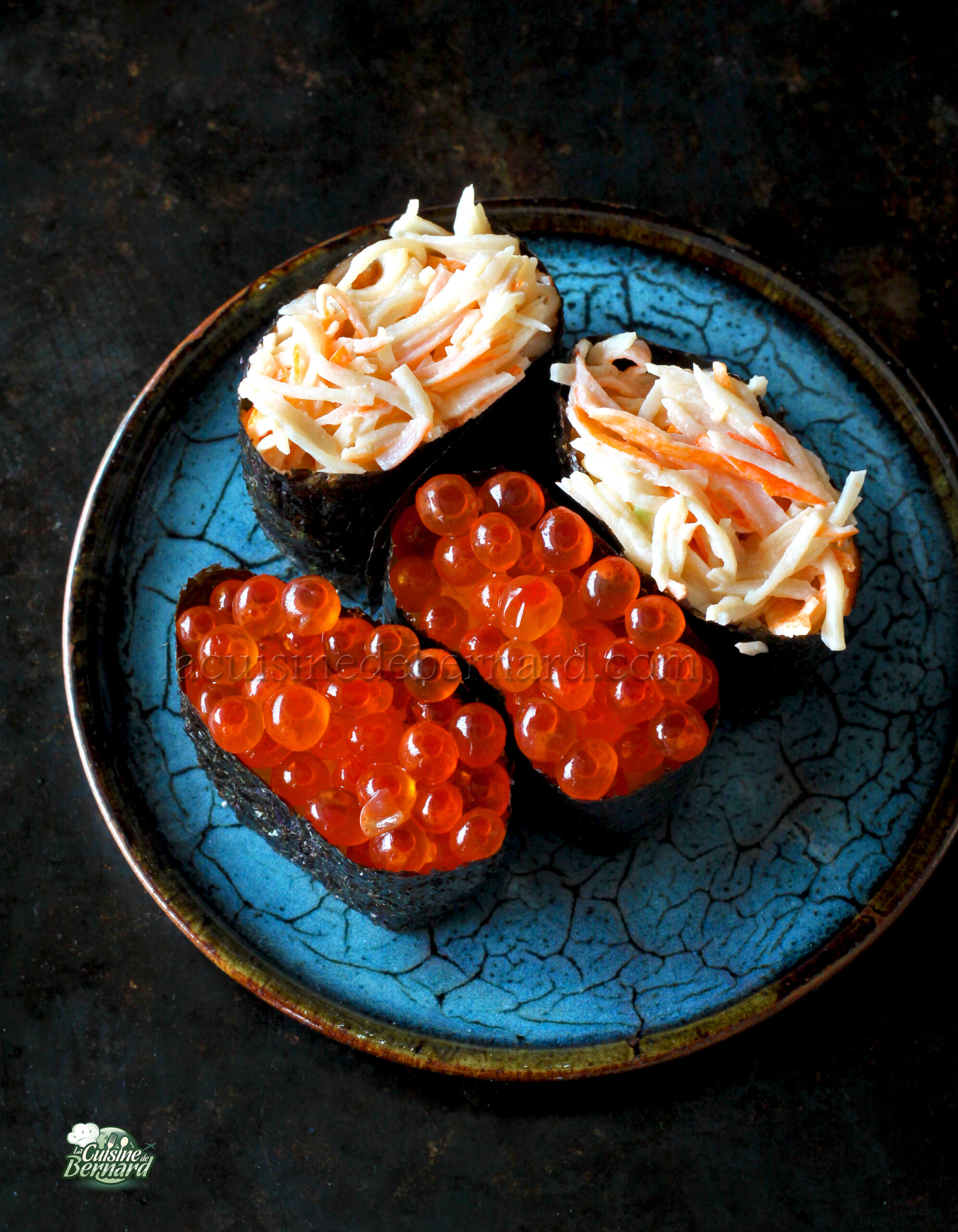 Une assiette bleue avec deux morceaux de sushi garnis de chair de crabe râpée et deux morceaux garnis d'œufs de poisson orange.