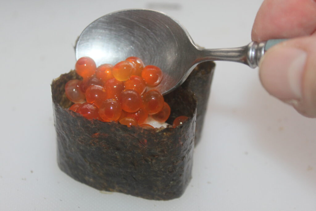Une cuillère dépose des œufs de poisson orange sur un morceau de sushi enveloppé dans des algues.