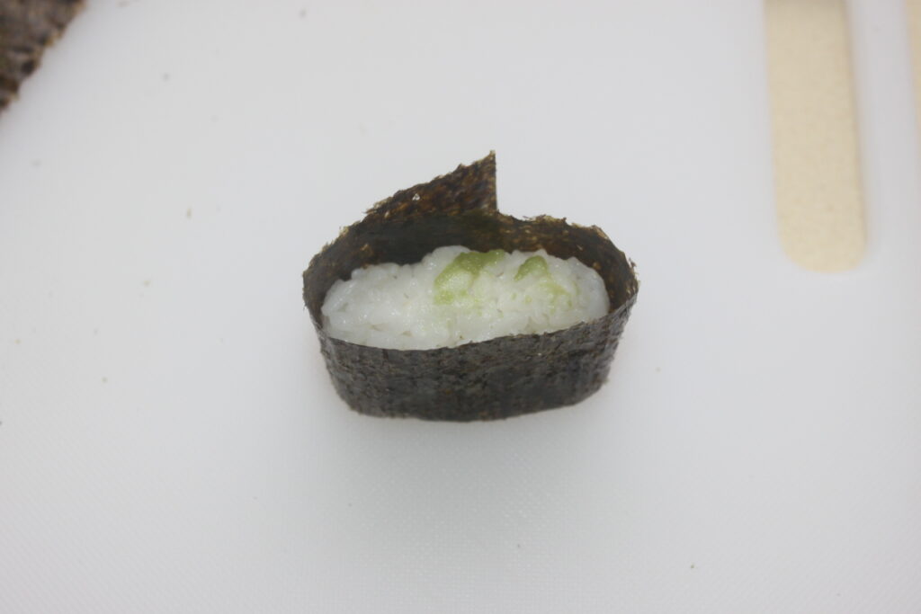 Sushi gunkan avec du riz blanc et une garniture verte enveloppés dans des algues sur une surface blanche.