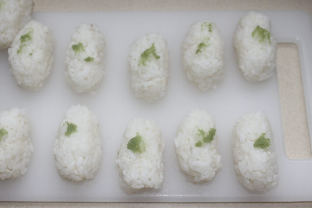 Dix boulettes de riz sur une planche à découper blanche, chacune garnie d'une petite quantité d'assaisonnement vert.