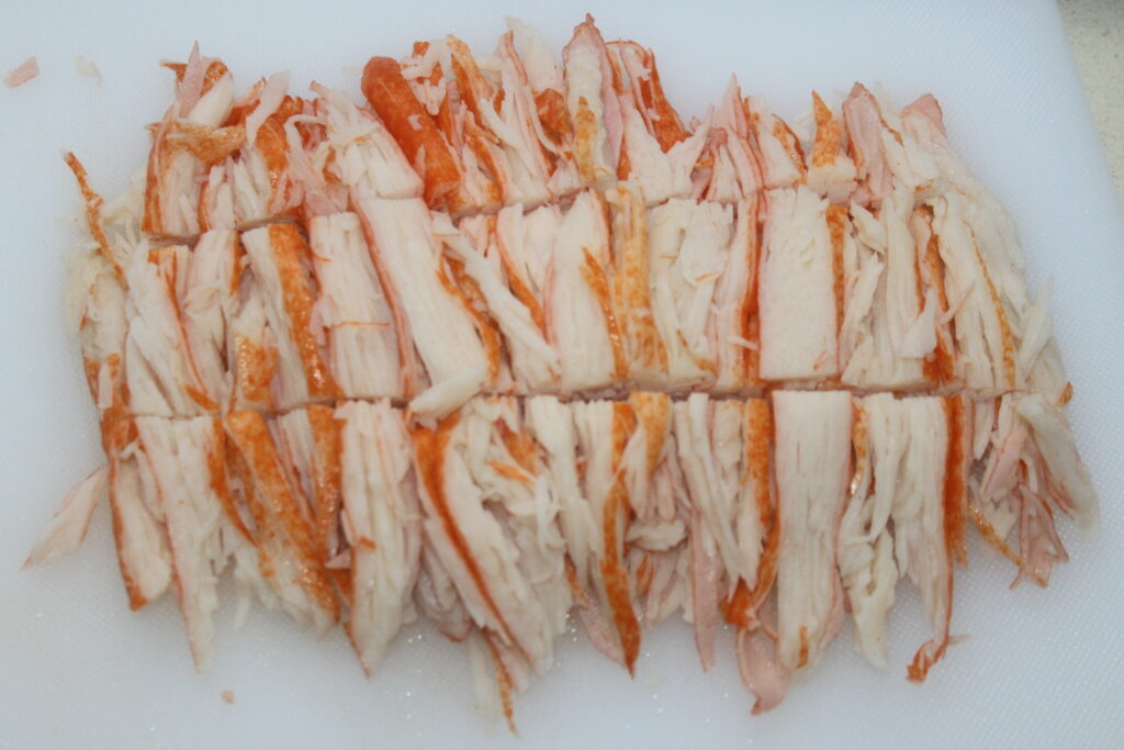 Des bâtonnets de crabe artificiels tranchés sont disposés soigneusement sur une planche à découper blanche.