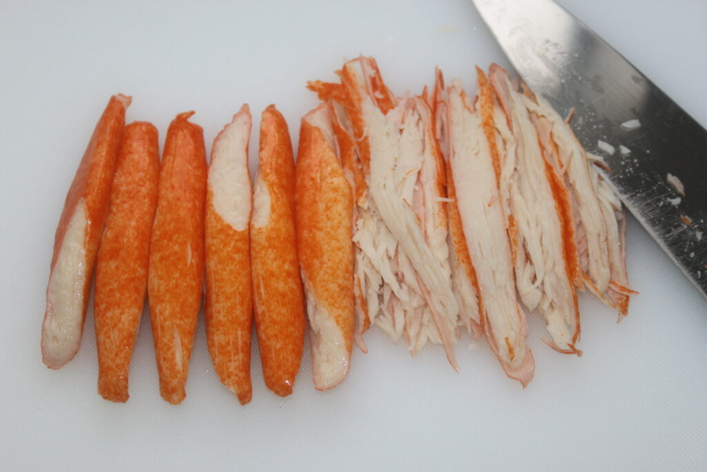 Bâtonnets de crabe artificiels, certains entiers et d'autres tranchés, sur une planche à découper à côté d'un couteau.