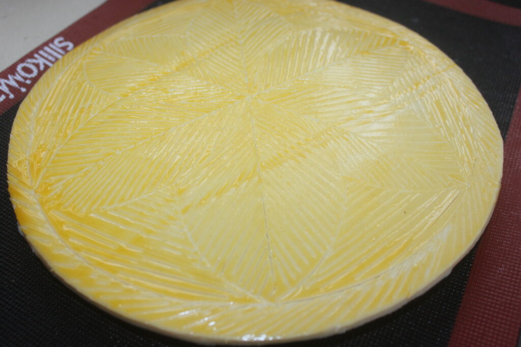 Une plaque ronde de beurre sur un tapis de cuisson en silicone, ornée d'un motif texturé en forme de feuille rappelant la surface d'une Galette des Rois.