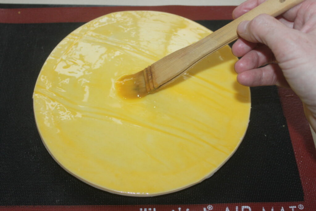 Une main utilise un pinceau pour appliquer une dorure à l'œuf sur une pâte à Galette des Rois ronde posée sur un tapis en silicone noir.