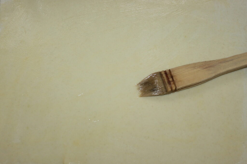 Gros plan d'un pinceau appliquant une couche translucide sur une surface beige clair, rappelant la brillance d'une Galette des Rois fraîchement cuite.