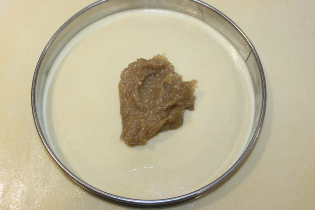 Un anneau métallique posé sur une surface plane contient une noisette de pâte brune, évoquant des cacahuètes onctueuses, posée sur une couche de substance blanche crémeuse.