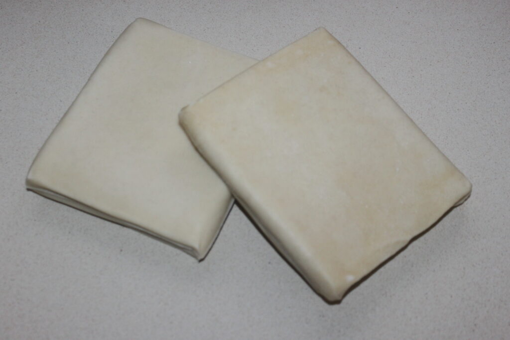 Deux morceaux carrés de pâte feuilletée crue reposent sur une surface plane, prêts à être transformés en une délicieuse Galette des Rois.