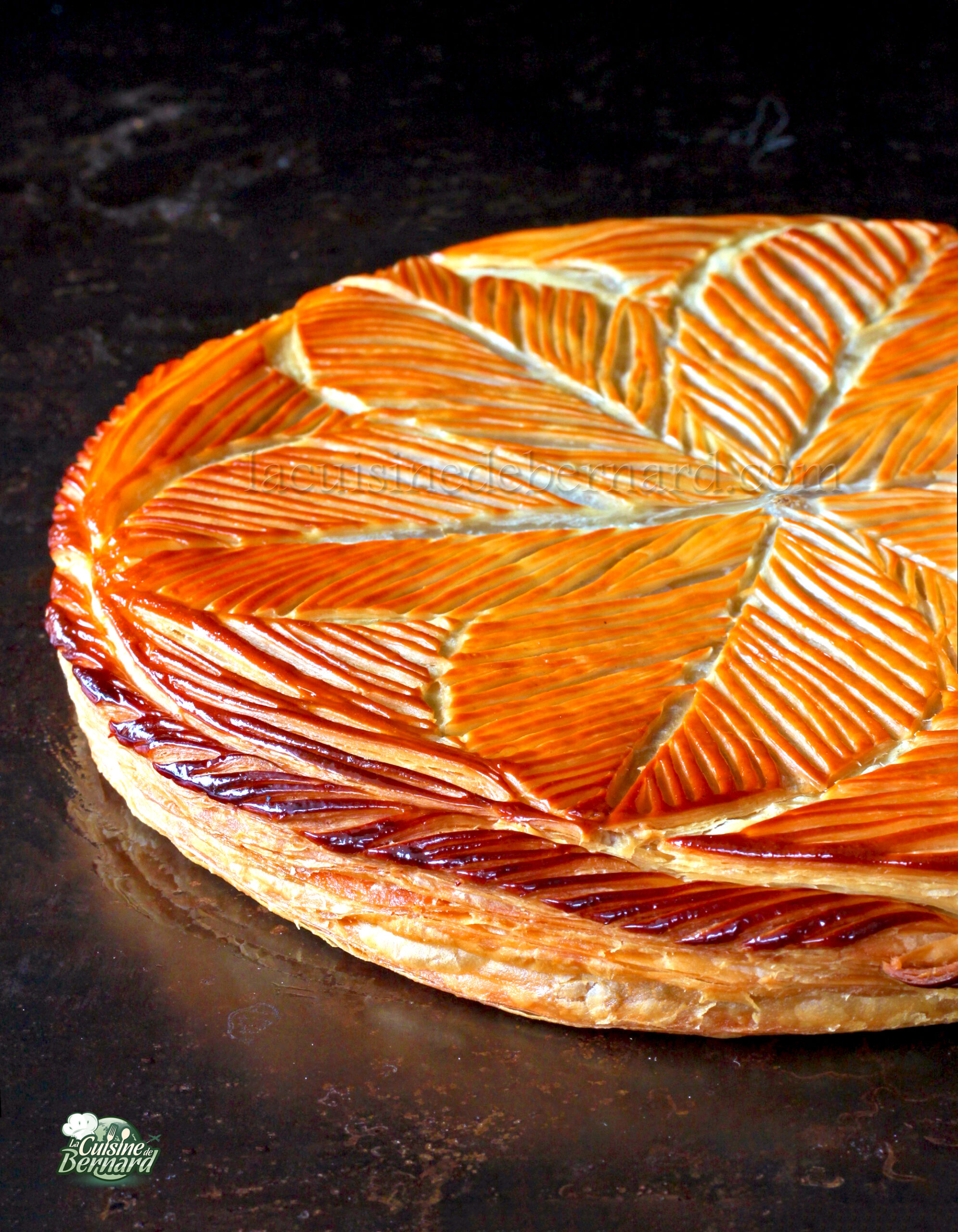 Une Galette des Rois feuilletée brun doré aux motifs de feuilles décoratives orne le fond sombre, son arôme évoquant une délicieuse touche de caramel.