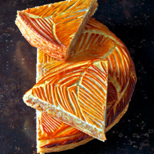 Une tarte ronde avec une croûte dorée au motif de feuilles complexes, partiellement tranchée pour révéler sa garniture en couches de caramel sur un fond sombre.