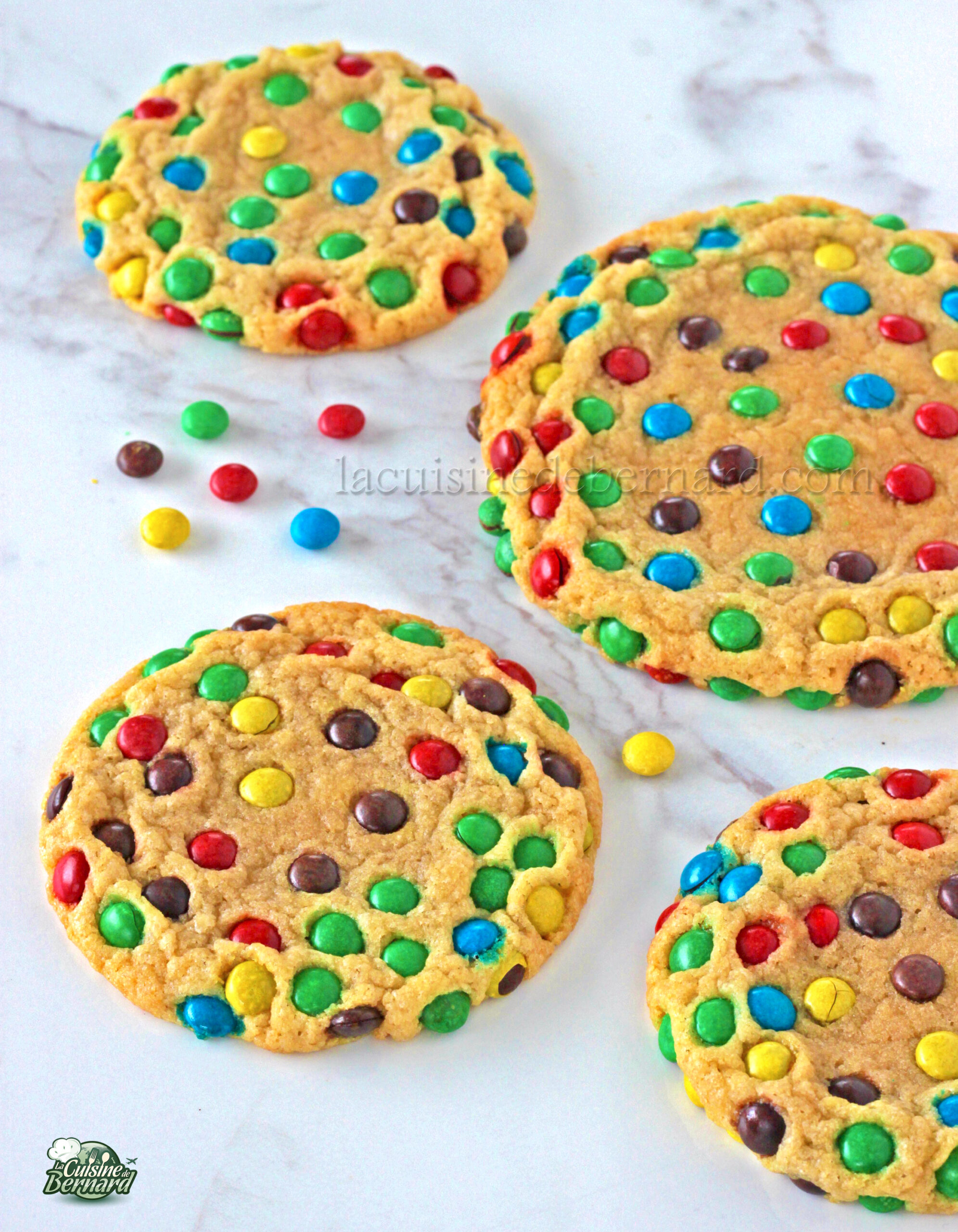 Cinq biscuits M&M's colorés, parsemés de mini M&M's vibrants, reposent de manière invitante sur une surface blanche, leurs bonbons dispersés ajoutant une touche de couleur supplémentaire.