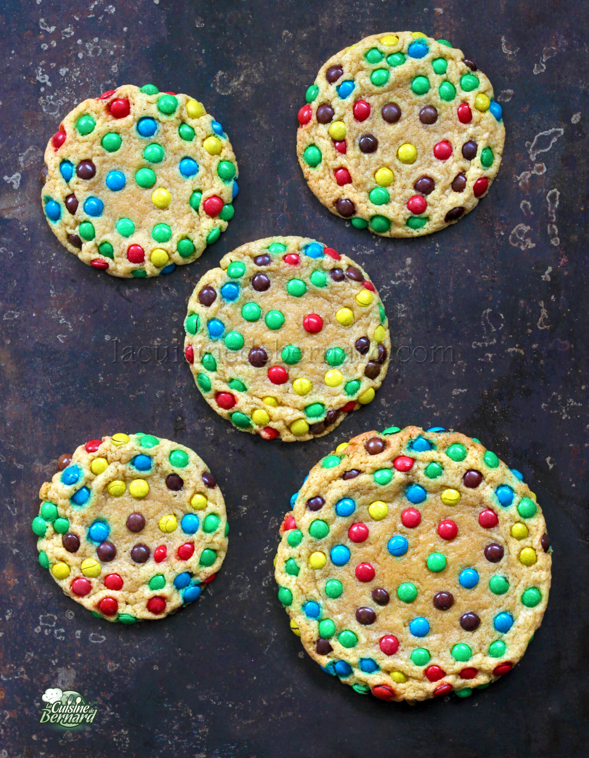 Cinq gros biscuits parsemés de mini M&M's vibrants sont astucieusement disposés sur une surface sombre, créant un contraste délicieux.