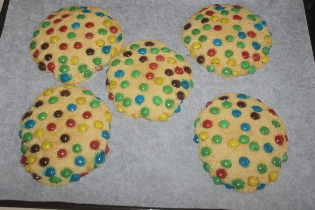 Cinq gros biscuits garnis de mini M&M's aux couleurs vives reposent sur une plaque à pâtisserie recouverte de papier sulfurisé.