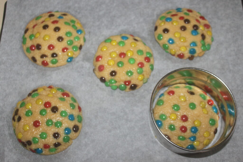 Cinq biscuits non cuits garnis de mini M&M's colorés sont soigneusement disposés sur une plaque à pâtisserie à côté d'un emporte pièce.