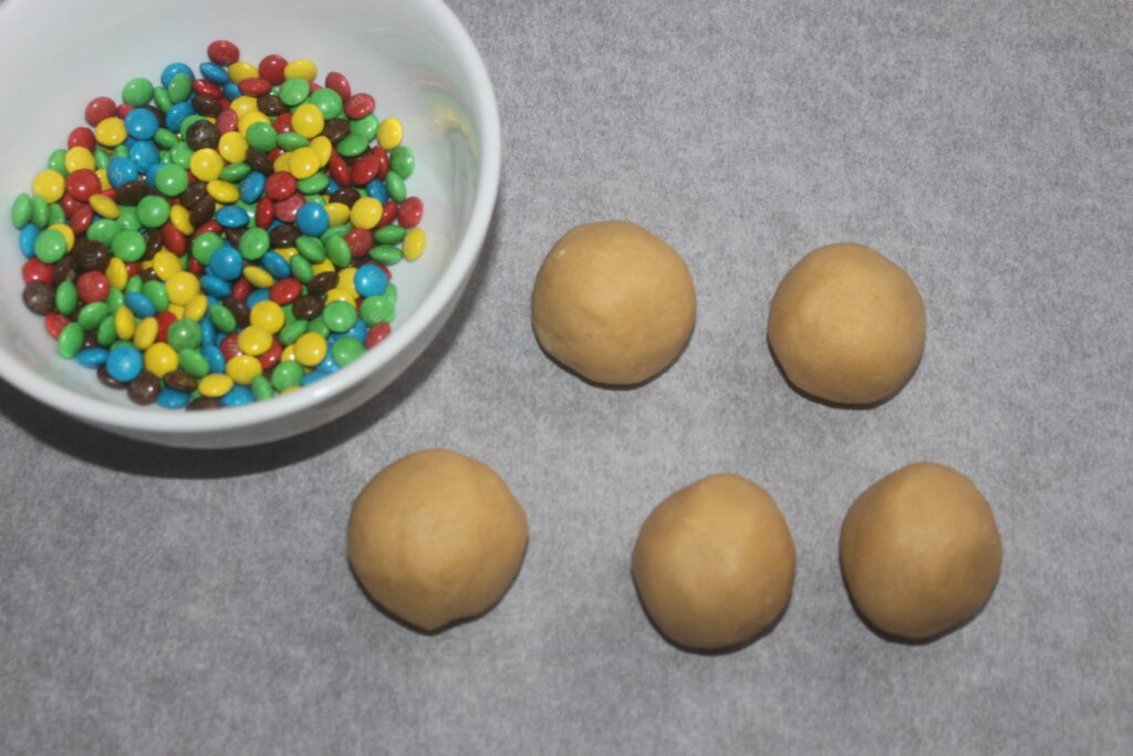 Un bol de mini bonbons au chocolat M&M's colorés est posé à côté de quatre boules de pâte à biscuits non cuites sur du papier sulfurisé.