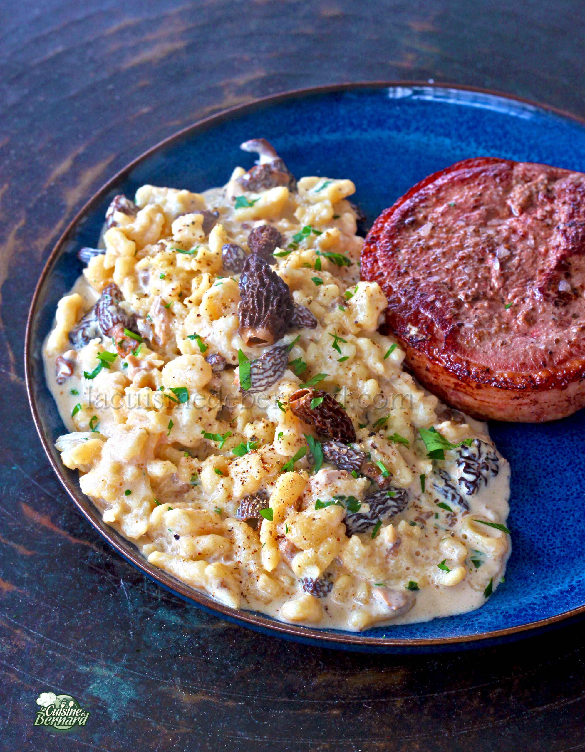 Une assiette composée de Spätzles crémeux aux morilles et aux herbes, accompagnés d'un magret grillé parfaitement doré, élégamment présenté sur un plat bleu.