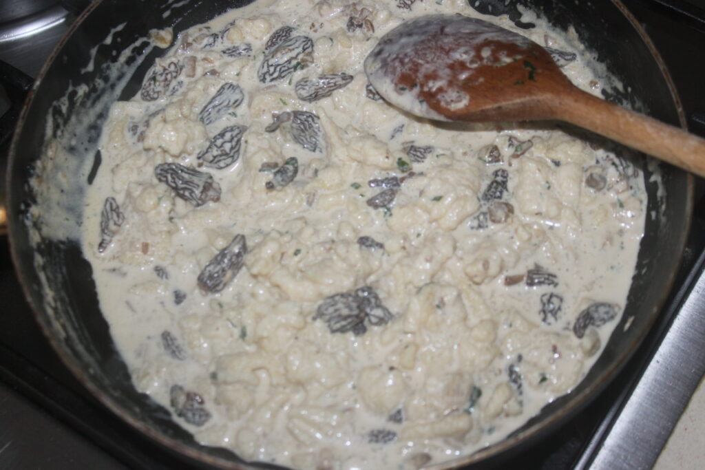 Une sauce crémeuse aux morilles est remuée dans une poêle avec une cuillère en bois, tandis que de délicats Spätzles attendent leur enrobage savoureux.