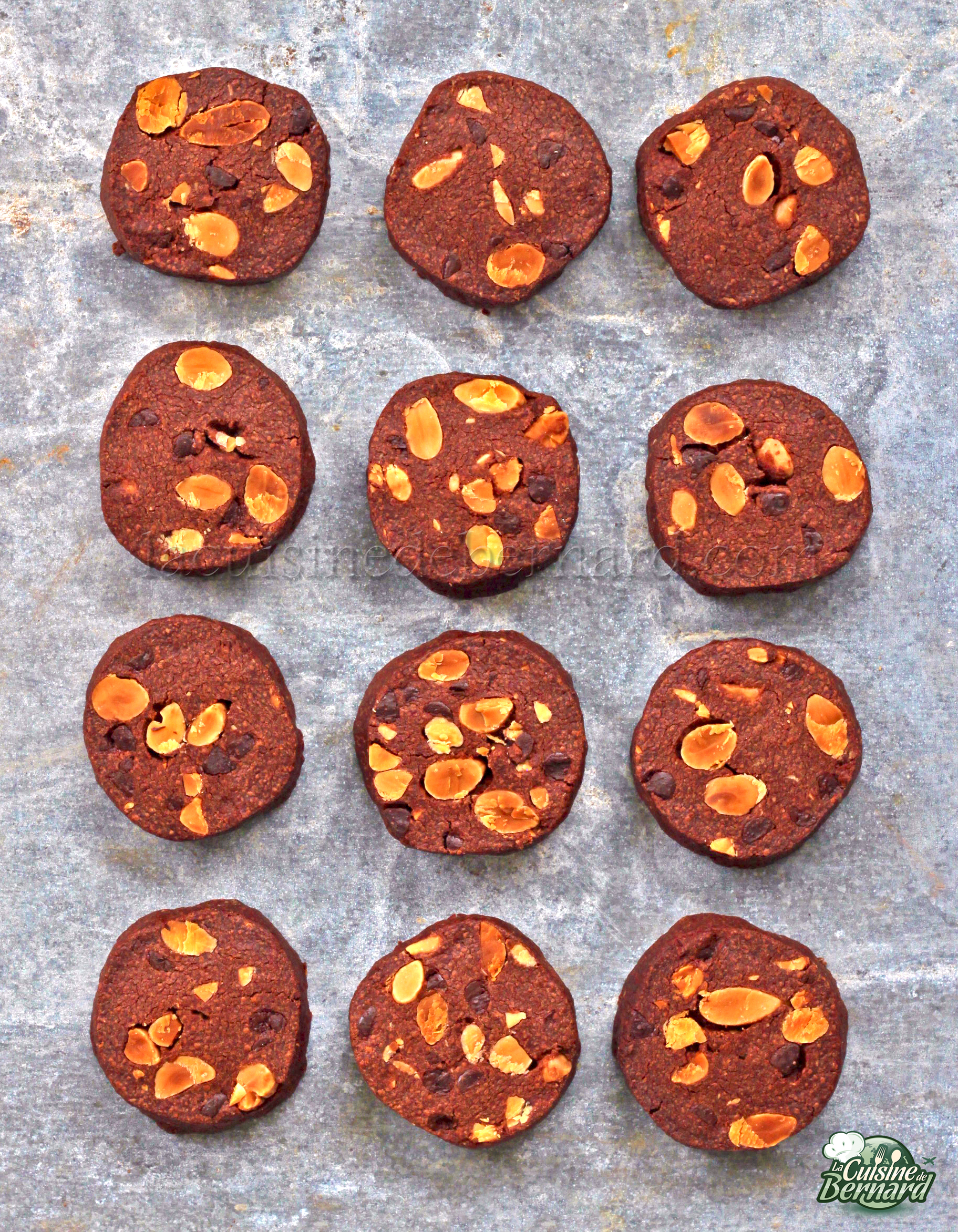 Douze sablés ronds au chocolat avec des tranches d'amandes sont disposés uniformément sur une surface grise texturée.