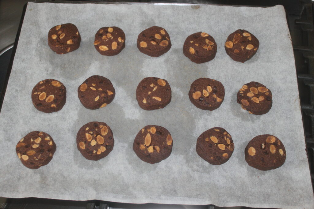 Quinze biscuits ronds au chocolat avec du cacao et des amandes grillées sont posés sur une plaque à pâtisserie recouverte de papier sulfurisé.