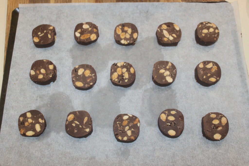 Seize tranches rondes de pâte à biscuits au chocolat avec amandes grillées, disposées en quatre rangées sur du papier sulfurisé.