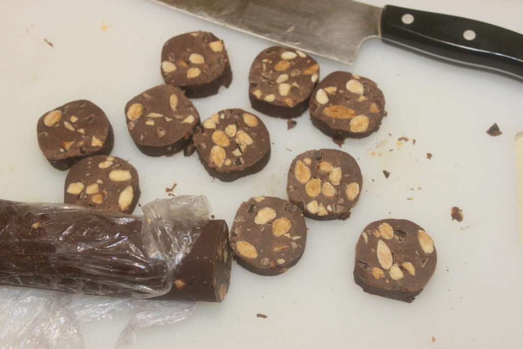 Un rouleau de chocolat et de noix coupé en tranches est posé sur une planche à découper avec un couteau. Certaines tranches sont encore attachées par une pellicule plastique, mettant en valeur la richesse du cacao et des amandes grillées.