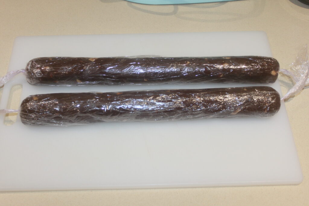 Deux rouleaux de salami au chocolat en forme de saucisse emballés dans du plastique reposent sur une planche à découper blanche, infusés de riches notes de cacao.