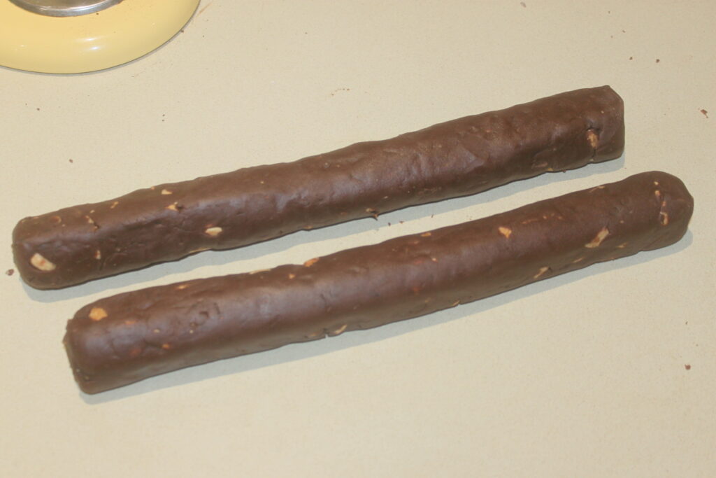 Deux bûches cylindriques de pâte riche en chocolat avec de petits morceaux de noix reposent de manière alléchante sur une surface beige, rappelant les sablés traditionnels.