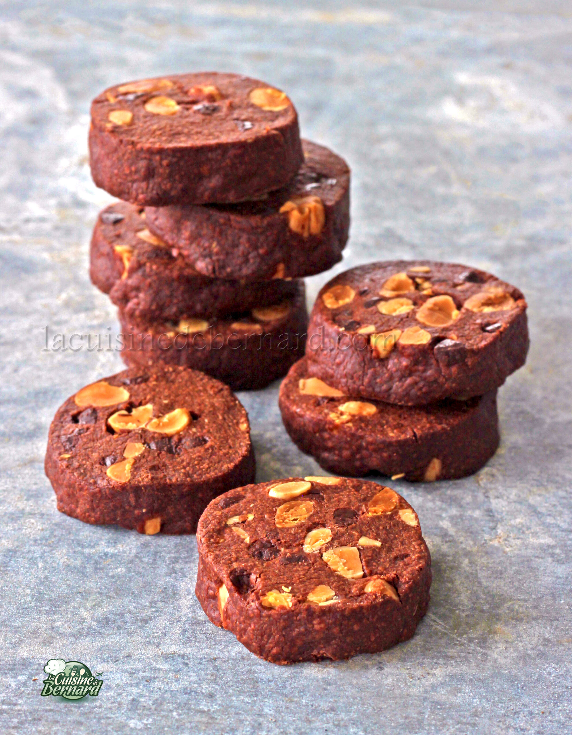 Une pile de sablés ronds au chocolat avec des noix visibles repose sur une surface grise texturée.