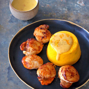 Des coquilles Saint Jacques grillées élégamment présentées sur une assiette noire, accompagnées de beurre à l'orange et d'un flan de potimarron vibrant. Une tasse de sauce supplémentaire trône en arrière plan, rehaussant l'attrait du plat.