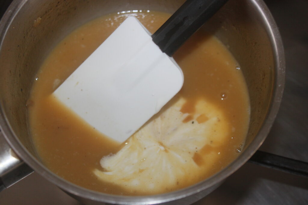 Une spatule repose dans une casserole contenant un liquide épais, brun orangé, avec une cuillerée de crème tourbillonnant dans le mélange, rappelant les riches saveurs d'un Flan de Potimarron.