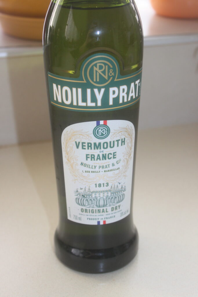 Une bouteille de Vermouth sec Original Noilly Prat de France, de 750 ml, avec une étiquette verte, orne une surface de couleur claire, parfaite pour accompagner les coquilles Saint Jacques.