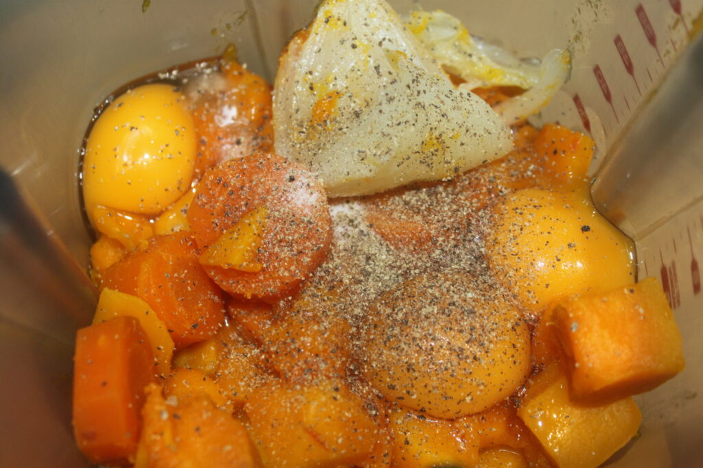 Un récipient de mixeur avec des œufs cassés, des morceaux de potimarron coupés, un citron pressé et du poivre noir saupoudré.