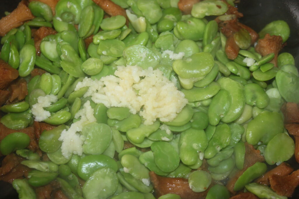 Un gros plan de fèves à l'ail émincé se mêle harmonieusement aux champignons cuits, évoquant le charme terreux des pâtes fèves girolles.