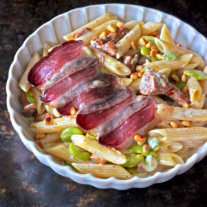 Un plat à base de penne, haricots verts, pignons de pin et tranches de magret fumé, élégamment présenté dans un plat rond blanc.
