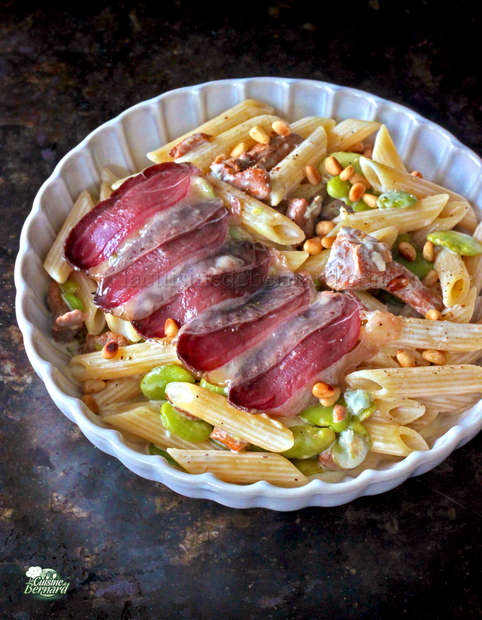 Un plat à base de penne, haricots verts, pignons de pin et tranches de magret fumé, élégamment présenté dans un plat rond blanc.