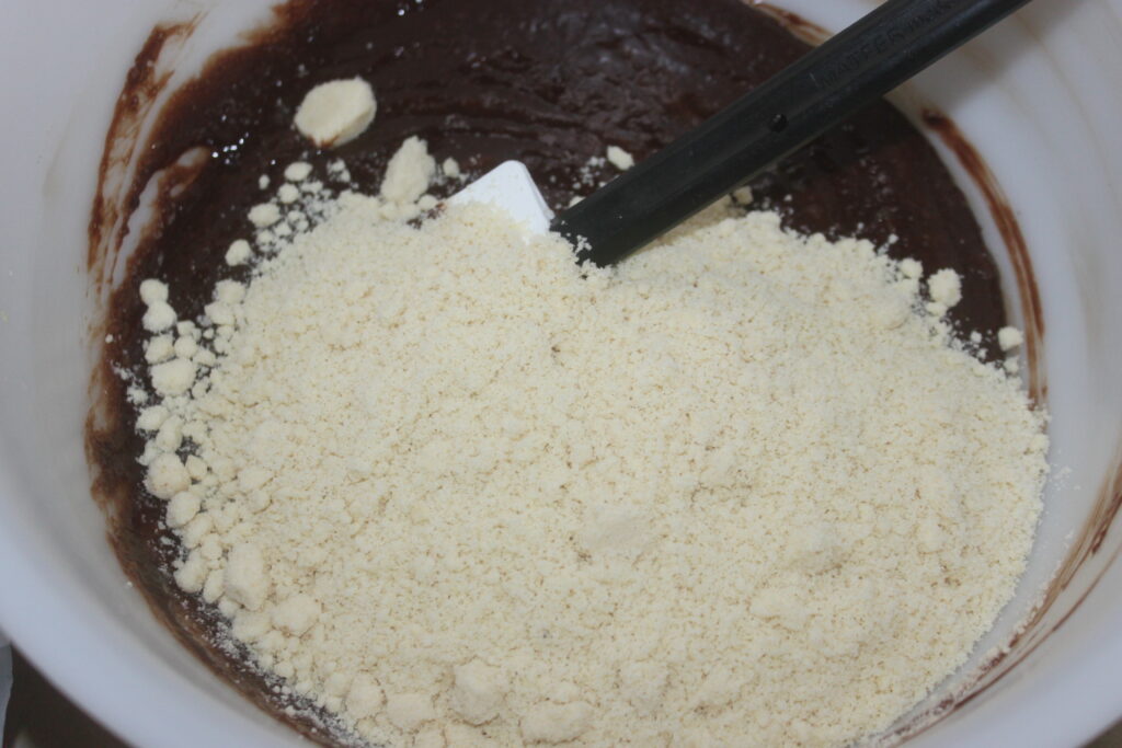 Un bol de pâte à brownie riche avec de la farine d'amande saupoudrée sur le dessus et une spatule reposant dans le mélange infusé de chocolat.